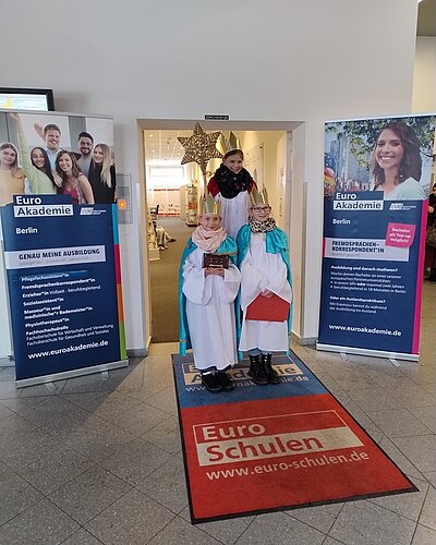 Im Eingangsbereich der Euro-Schulen und der Euro Akademie Berlin stehen drei als Sternsinger verkleidete Kinder in weißen Gewändern mit farbigen Umhängen und goldenen Kronen. Sie halten ein Buch und einen Sternstab. Im Hintergrund ist ein großer goldener Stern über der Tür angebracht. Links und rechts des Eingangs stehen zwei Roll-Ups mit den Logos und Informationen der Euro Akademie Berlin. Auf dem Boden liegt eine rote Fußmatte mit den Logos der Euro-Schulen und der Euro Akademie. Die Szene ist hell ausgeleuchtet und vermittelt eine festliche Atmosphäre am 6. Januar 2026.
