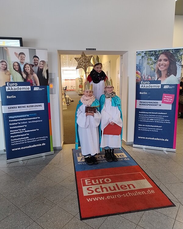 Im Eingangsbereich der Euro-Schulen und der Euro Akademie Berlin stehen drei als Sternsinger verkleidete Kinder in weißen Gewändern mit farbigen Umhängen und goldenen Kronen. Sie halten ein Buch und einen Sternstab. Im Hintergrund ist ein großer goldener Stern über der Tür angebracht. Links und rechts des Eingangs stehen zwei Roll-Ups mit den Logos und Informationen der Euro Akademie Berlin. Auf dem Boden liegt eine rote Fußmatte mit den Logos der Euro-Schulen und der Euro Akademie. Die Szene ist hell ausgeleuchtet und vermittelt eine festliche Atmosphäre am 6. Januar 2026.