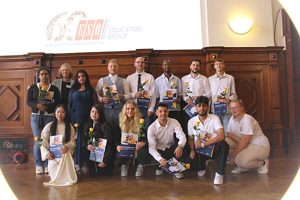 Gruppenbild mit festlich gekleideten Menschen. Die meisten Menschen halten Blumen und Zeugnismappen in den Händen. Im Hintergrund ist über einer holzvertäfelten Wand das 60-Jahre-Logo der ESO Education Group an die Wand projiziert.