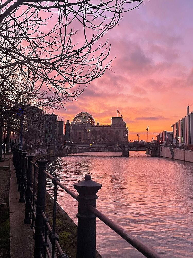 Abendhimmel über Berlin. Im Zentrum des Bildes erkennt man im Hintergrund das Reichstagsgebäude, an dem die Spree entlangfließt. Die Sonne geht unter und färbt den Himmel in warmen Orange- und Rosatönen, der sich im Wasser spiegelt. Im Vordergrund sind kahlen Bäume und ein Metallgeländer zu sehen.