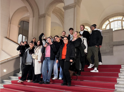 Eine Gruppe von jungen Menschen posiert lächelnd auf einer breiten Treppe mit rotem Teppichboden in einem historischen Gebäude mit hohen, gewölbten Decken und großen Fenstern.