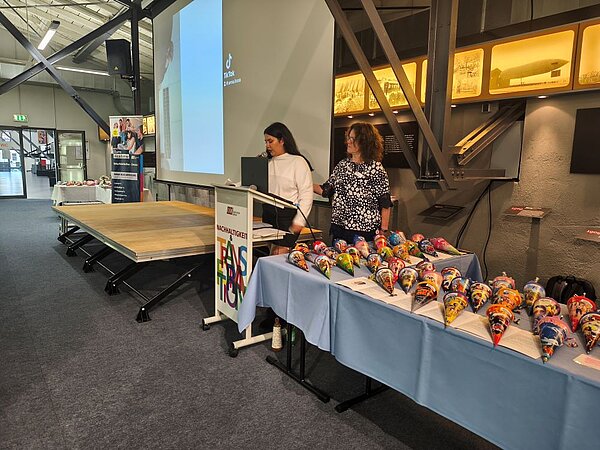 Herzlicher Abschied, freudiger Anfang: Pflege-Absolvent*innen geehrt und neue Auszubildende willkommen geheißen Zwei Frauen stehen an einem Podium neben einem Tisch, der mit vielen bunten, kleinen Zuckertüten bedeckt ist. Eine der Frauen spricht ins Mikrofon, während die andere zuhört. Im Hintergrund ist eine Leinwand mit einer Präsentation zu sehen.