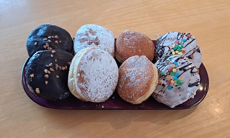 Sechs verschiedene Berliner (Pfannkuchen, Krapfen, Kreppel) auf einem länglichen, dunklen Teller: zwei mit Schokoladenglasur und Nüssen, zwei mit Puderzucker bestäubt, einer mit einfacher Zuckerglasur und einer mit bunter Streuselglasur.