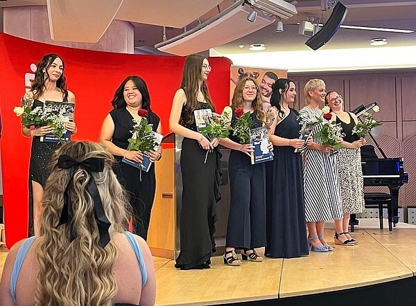 Feierlicher Abschied: Exmatrikulation der Absolvent*innen 2025 Eine Gruppe von Personen steht auf einer Bühne und hält Blumen und Zertifikate. Sie sind elegant gekleidet und lächeln. Im Hintergrund ist ein rotes Banner und ein Klavier zu sehen.