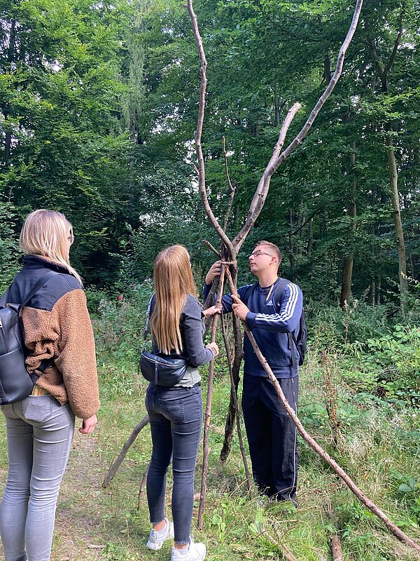 Kennenlerntag der Euro Akademie Rochlitz Drei Jugendliche bauen im Wald eine Konstruktion aus langen Ästen, die wie ein Tipi aufgestellt werden.
