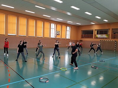 In einer Turnhalle mit großen Fenstern und heruntergelassenen Jalousien stehen Schülerinnen einen Lehrer gegenüber. Sie stehen im Ausfallschritt und strecken dabei einen Arm vor der Brust nach vorne. Auf dem Boden liegen Übungsgeräte, vor der Wand steht ein Handballtor.