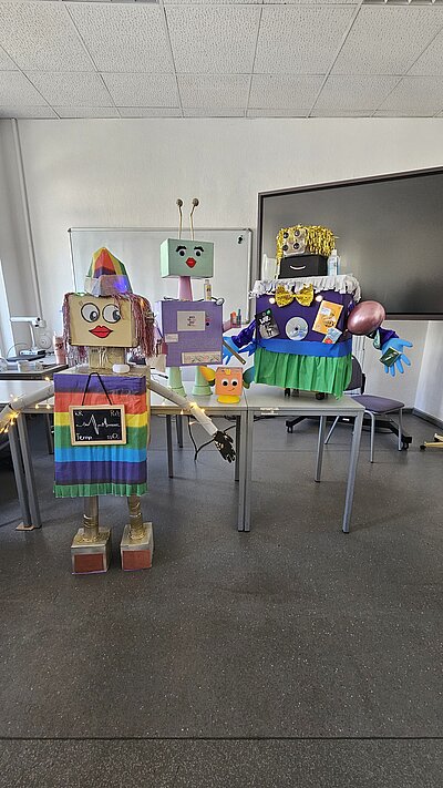 Die vier Roboter präsentieren sich: Zwei kleine und ein Mini-Roboter stehen auf den Tischen, ein großer Roboter steht davor auf dem Boden. Sie bestehen aus bunt bemalten Kartons, buntem Krepppapier, Lametta-Haaren, Händen aus Plastikhandschuhen und sind mit Luftballons und CDs dekoriert.