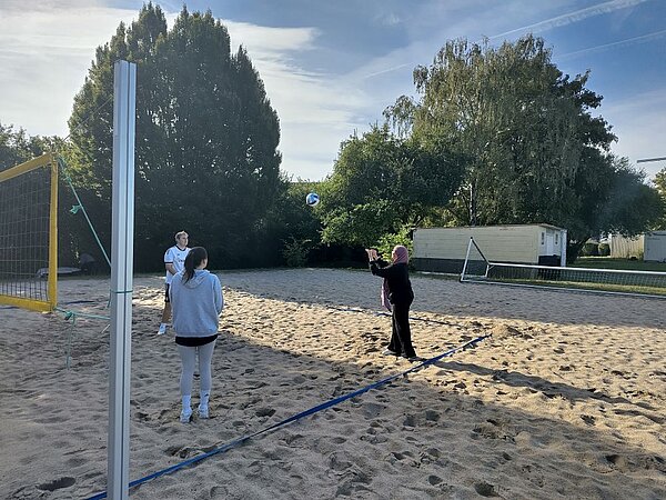Drei Personen spielen Beachvolleyball auf einem Sandplatz. Im Hintergrund sind hohe Bäume und ein sonniger Himmel zu erkennen.