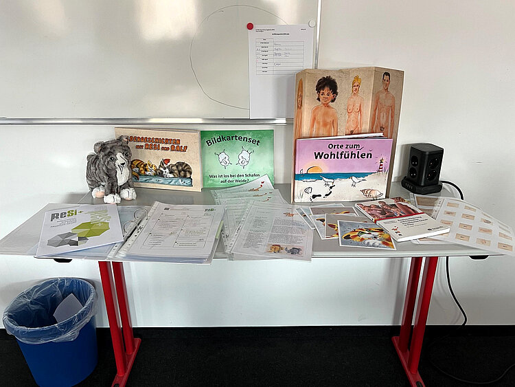 Wo die Katze nicht gestreichelt werden will Auf einem Tisch sind verschiedene Medien zum Projekt versammelt: die Broschüre "ReSi+", das Buch "Bildergeschichten mit Resi und Ralf", ein Bildkartenset namenes "Was ist los bei den Schafen auf der Weide", das Buch "Orte zum Wohlfühlen" und ein Ordner mit verschiedenen Arbeitsblättern.