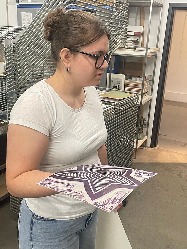 Eine Person mit Brille und Dutt hält ein großformatiges, violett-weißes Kunstwerk, das einen Stern darstellt, in den Händen. Sie trägt ein weißes T-Shirt und eine blaue Jeans. Im Hintergrund sind eine Metallkonstruktion und Metallregale mit Büchern zu sehen.