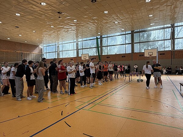 In einer Sporthalle mit gelbem Boden und hohen Fenstern stehen mehrere Gruppen junger Menschen in Sportkleidung in einer Reihe. Zwei Personen im Vordergrund reichen sich die Hand, während andere zusehen. Im Hintergrund sind Basketballkörbe und Volleyballnetze zu erkennen. Die Szene wirkt wie eine Siegerehrung oder ein Abschluss eines Sportturniers.