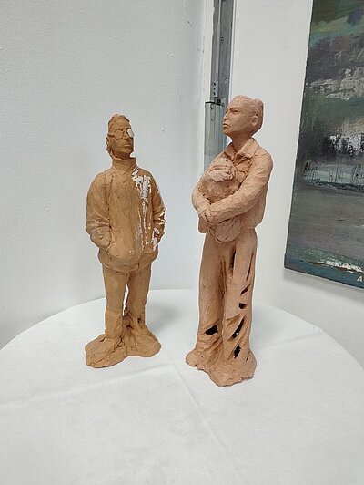 Zwei Tonfiguren in orange-braunen Tönen, die Menschen darstellen, stehen auf weißer Oberfläche in einem Ausstellungsraum. Im Hintergrund ein abstraktes Gemälde in Grün-, Weiß- und Grautönen.