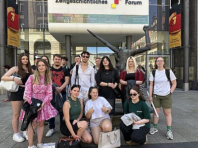 Eine Gruppe Jugendlicher steht vor dem Eingang des Zeitgeschichtlichen Forums in Leipzig. Im Hintergrund ist das Gebäude mit Schriftzug „Zeitgeschichtliches Forum“ sowie die Fahnen zur Ausstellung „Unsere Geschichte“ zu sehen.