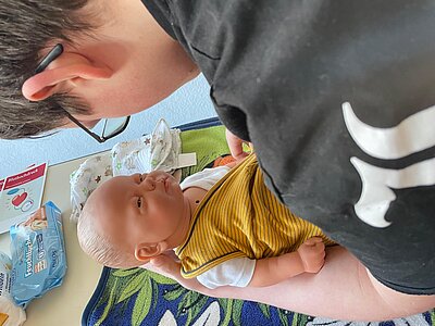 Ein junger Mann hält eine Babypuppe; auf dem Tisch vor ihm sind Wickelutensilien zu sehen.