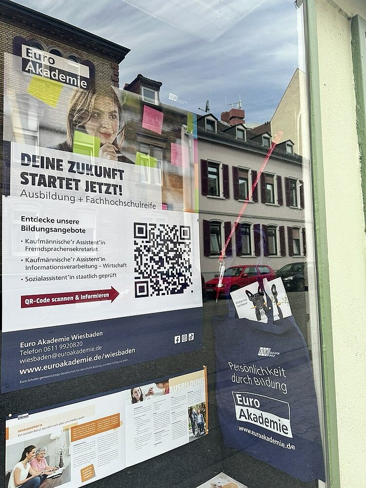 Plakat mit Werbemotiv für die Euro Akademie Wiesbaden, das an einer Fensterscheibe angebracht ist. Im oberen Bereich ist ein Porträt einer Person mit langem Haar zu sehen. Darunter steht ein großflächiger Text, daneben ein QR-Code. Im unteren Bereich sind weitere Informationen und Kontaktdaten abgedruckt. Rechts unten hängt eine dunkelblaue Stofftasche mit dem Logo der Euro Akademie. Im Hintergrund spiegeln sich Fassaden von Wohnhäusern mit weißen Fensterrahmen und roten Dächern.