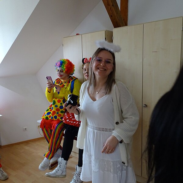 Eine Frau in einem weißen Kleid mit Engelsflügeln und einem Heiligenschein lächelt in die Kamera. Hinter ihr sitzen zwei als Clowns verkleidete Personen auf einer Bank, eine von ihnen tippt auf ihrem Handy.