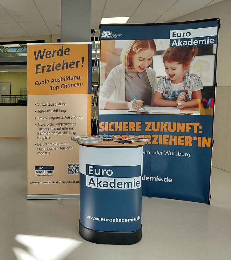 Ein Infotisch der Euro Akademie mit einem Roll-up-Banner im Hintergrund, das den Slogan „Sichere Zukunft: Erzieherin“ und „Werde Erzieher! Coole Ausbildung – Top Chancen“ zeigt. Auf dem Tisch liegen Flyer und Informationsmaterialien.