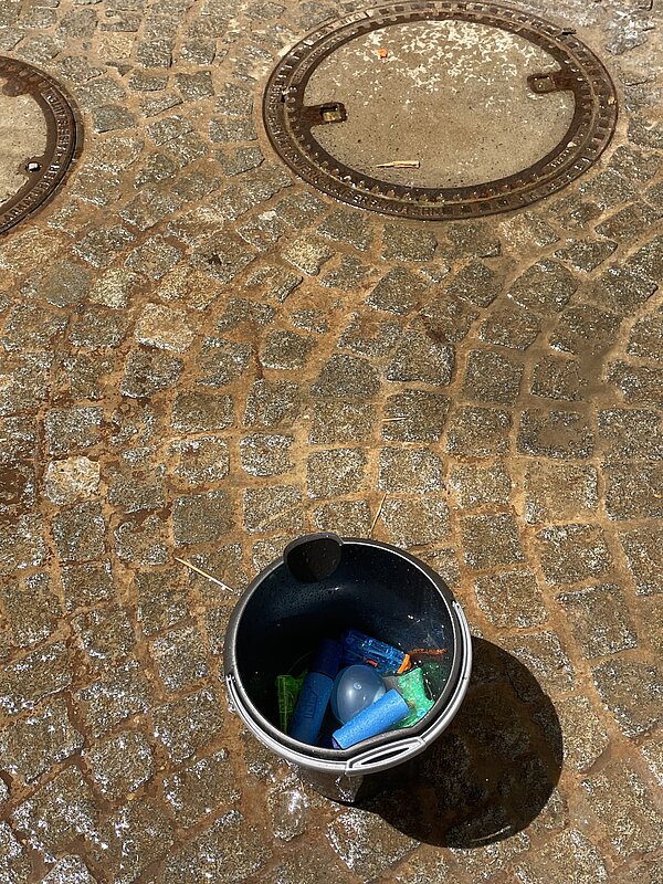 Ein schwarzer Eimer steht auf einem nassen Kopfsteinpflaster. Darin befinden sich bunte Wasserpistolen und Wasserbomben. Im Hintergrund sind zwei Kanaldeckel zu sehen.
