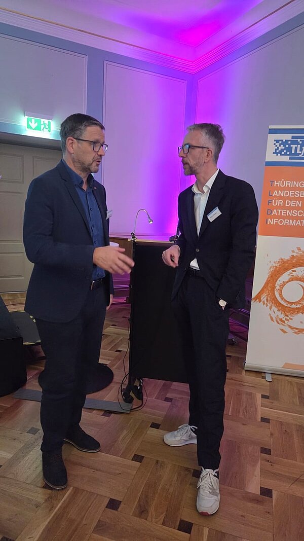 Leiter Uwe Schöneck und ein Referent stehen zusammen und unterhalten sich. Beide tragen einen dunklen Anzug und Sneaker.