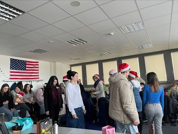 Eine Gruppe von Menschen in einem Klassenraum, die Weihnachtsmützen tragen und sich unter einem amerikanischen Flaggenbanner versammeln. Einige Personen halten Geschenke oder Tüten, andere laufen im Kreis herum. Die Atmosphäre scheint fröhlich und festlich zu sein.