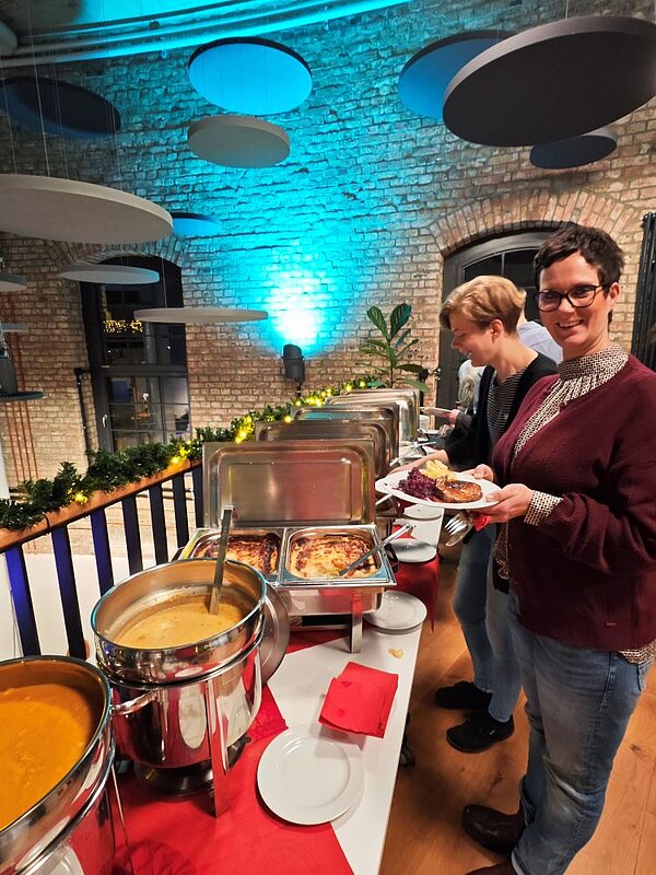 Zwei lächelnde Personen stehen an einem Buffet-Tisch in einem Raum mit Ziegelsteinwänden. Eine Person hält einen Teller mit Essen in der Hand. Auf dem Tisch sind silberne Warmhaltebehälter mit verschiedenen Speisen, wie z.B. Nudelgerichten, und eine große Schüssel mit Suppe. Der Tisch ist mit einer roten Tischdecke bedeckt.
