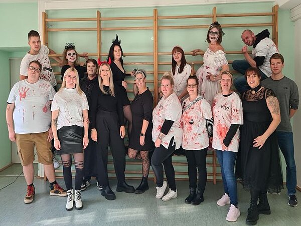 Eine Gruppe in Halloween-Outfits hat sich für das Foto in einem Turnraum vor einem Klettergerüst positioniert. Manche Personen tragen schwarze Kleidung, andere haben auf weiße T-Shirts rote Farbe gespritzt, die wie Blut aussieht.