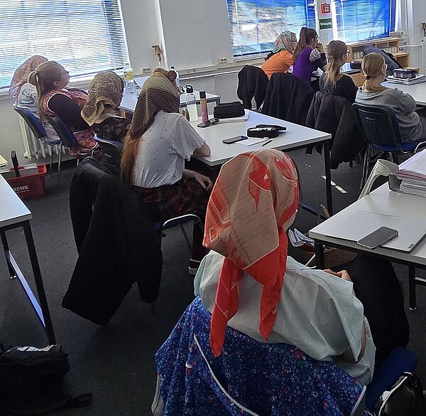 Schülerinnen sitzen in einem Klassenraum, alle mit Kopftuch und Schürze verkleidet, vermutlich passend zum Motto „Rentner-Tag“. Sie schauen nach vorn in Richtung Lehrkraft.
