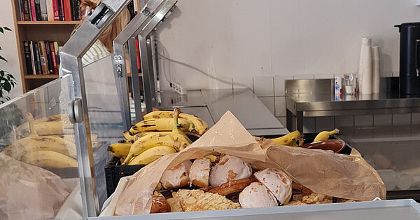 Essensausgabe mit einer Kiste Bananen, belegten Brötchen und Brezeln unter einer Plexiglasabdeckung.