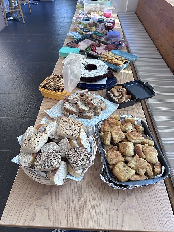 Gemeinsam essen. Lachen. Starten! Ein langer Tisch mit einer Auswahl an Speisen und Lebensmitteln. Im Vordergrund befinden sich in Schalen verschiedene Gebäckstücke wie Brötchen und Kuchen.