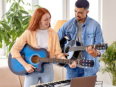 Ein Mann und eine Frau stehen in einem hellen Wohnzimmer und spielen Gitarre. Die Frau links hält eine blaue Gitarre und lächelt glücklich, während sie spielt. Der Mann rechts trägt ein blaues Hemd und spielt ebenfalls Gitarre, wobei er sich auf sein Instrument konzentriert. Vor ihnen steht ein Keyboard auf einem Tisch, auf dem auch ein Laptop geöffnet ist. Im Hintergrund sind große Fenster und Zimmerpflanzen zu sehen, die dem Raum eine freundliche und entspannte Atmosphäre verleihen. Beide wirken fröhlich und engagiert.