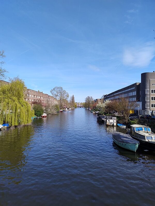 Ein ruhiger Wasserweg mit kleinen Booten, die an einem Steg festgemacht sind. Auf der linken Seite stehen moderne Wohnhäuser, und der Himmel ist klar und blau.