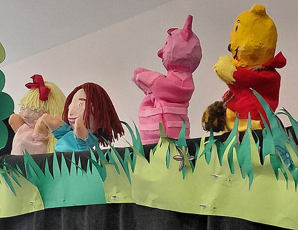 Vier Handpuppen mit unterschiedlichen Farben und Formen stehen hinter einer Kulisse aus grünem Papier, das als Gras gestaltet ist. Die Puppen zeigen verschiedene Figuren, darunter eine gelbe Figur mit rotem Oberteil, eine rosa gestreifte Figur, eine Puppe mit langen braunen Haaren und eine weitere mit blonden Haaren und roter Schleife. Die Szene wirkt wie eine Aufführung auf einer Puppenbühne.