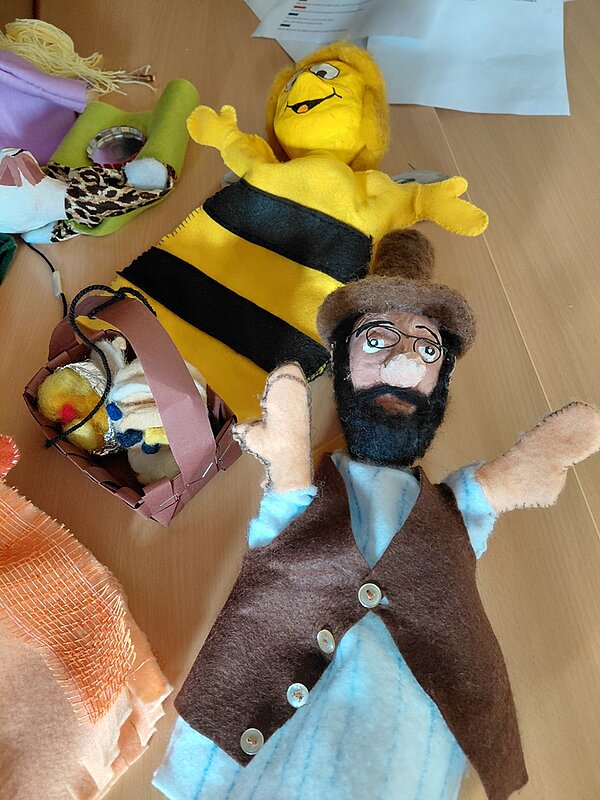 Im Vordergrund liegt eine Stoffpuppe mit brauner Weste, hellblauem Hemd, dunklem Bart und Brille. Daneben befindet sich eine gelb-schwarz gestreifte Stofffigur mit breitem Lächeln und ausgestreckten Armen. Im Hintergrund sind weitere Stofftiere und ein kleiner Korb mit Plüschtieren zu sehen. Die Szene spielt auf einer hellen Holzoberfläche.