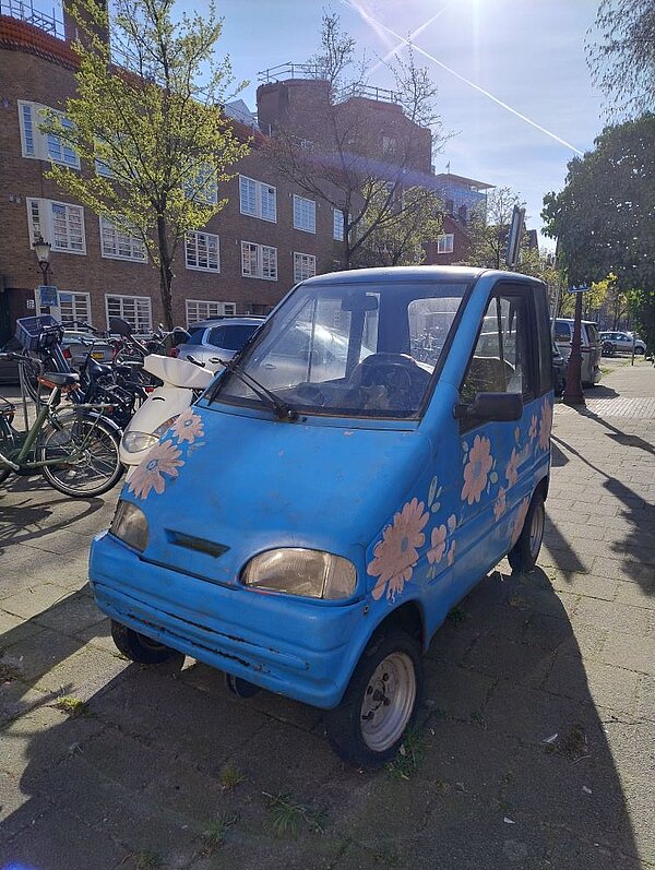Ein altes, blaues Auto mit Blumenmustern auf der Motorhaube, das auf einem Gehweg in einer Stadt geparkt ist. Im Hintergrund sind ein mehrstöckiges Wohnhaus und Fahrräder zu erkennen.