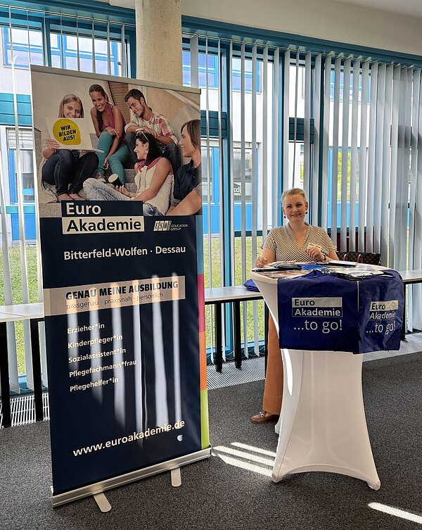 Großer Andrang bei der „Nacht der Berufe“ Eine Frau steht an einem Tisch mit Informationsmaterialien, die mit der Euro Akademie in Bitterfeld-Wolfen und Dessau zu tun haben. Hinter ihr steht ein Roll-up-Banner, das verschiedene Ausbildungsberufe wie Erzieher*in, Kinderpfleger*in, Sozialassistent*in, Pflegefachmann/-frau und Pflegehelfer*in bewirbt.