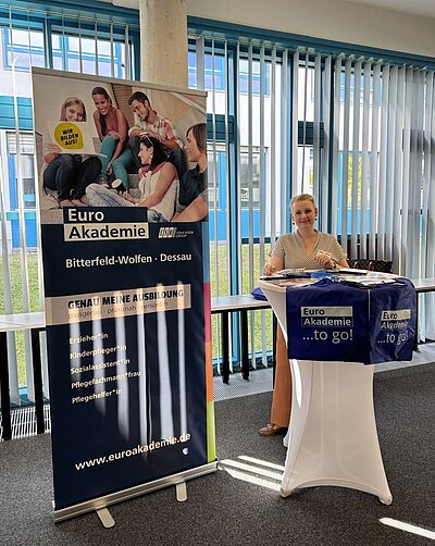 Eine Frau steht an einem Tisch mit Informationsmaterialien, die mit der Euro Akademie in Bitterfeld-Wolfen und Dessau zu tun haben. Hinter ihr steht ein Roll-up-Banner, das verschiedene Ausbildungsberufe wie Erzieher*in, Kinderpfleger*in, Sozialassistent*in, Pflegefachmann/-frau und Pflegehelfer*in bewirbt.