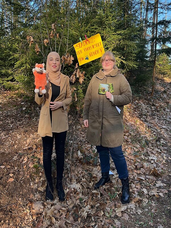 Zwei junge Frauen stehen vor einem Nadelbaum, an dem ein gelbes Schild mit der Aufschrift: "Station 2: Der Fuchs testet sein Gehör". Die Personen halten eine Fuchshandpuppe und Karten in der Hand.