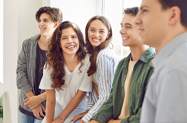 Jetzt bewerben! Eine Gruppe fünf Jugendlichen lehnen oder sitzen an einem Fenster und lächeln. Zwei junge Frauen davon schauen direkt in die Fotokamera, drei junge Männer sind im Profil zu sehen.