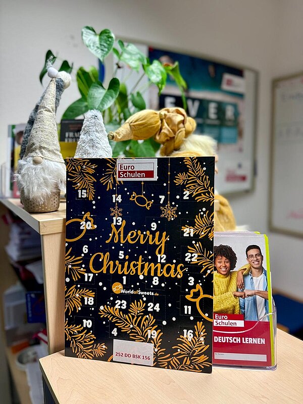 Ein weihnachtlich dekorierter Raum der Euro-Schulen und Euro Akademien in Dresden, Meißen und Riesa zeigt einen Adventskalender mit der Aufschrift 'Merry Christmas' in goldener Schrift auf schwarzem Hintergrund, verziert mit goldfarbenen Tannenzweigen und Sternen. Der Adventskalender trägt das Logo der Euro-Schulen und ist auf einem hellen Holztisch platziert. Daneben steht ein kleiner Aufsteller mit dem Titel 'Deutsch lernen' und einem Bild von zwei Personen, die lächelnd in die Kamera blicken. Im Hintergrund sind grüne Pflanzen und weitere Dekorationen zu erkennen, die eine festliche Atmosphäre schaffen. Vor dem Adventskalender stehen drei kleine, handgefertigte Weihnachtswichtel aus Stoff mit langen Zipfelmützen in Grau- und Beigetönen. Die gesamte Szene vermittelt eine warme, einladende Weihnachtsstimmung in den Räumlichkeiten der Bildungseinrichtungen.