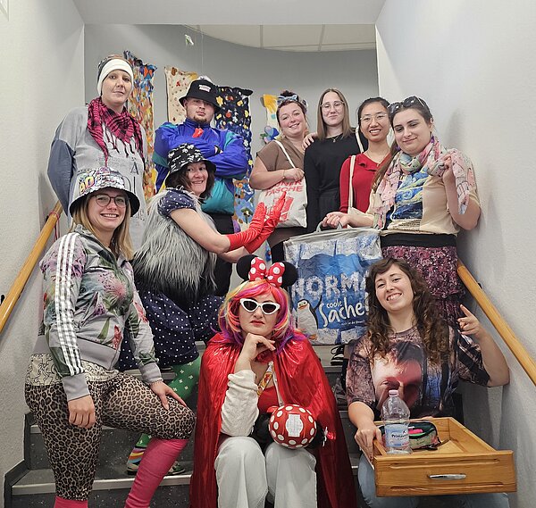 Die letzte Schulwoche der PFM 22: Ein unvergesslicher Abschluss Eine weitere Gruppe steht auf einer Treppe, diesmal in sehr bunten und auffälligen Kostümen – darunter ein Minnie-Maus-Outfit, ein Jogginganzug, bunte Hüte und schrille Accessoires. Eine Person hält ein Holztablett mit Wasserflasche und Requisiten.