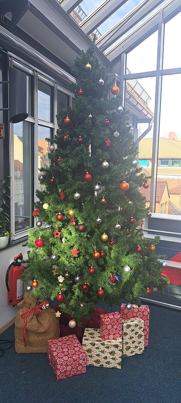 Ein hochgewachsener, künstlicher Weihnachtsbaum steht in einem hellen, modernen Raum mit großen Fenstern und sichtbarem Stahlrahmen. Der Baum ist reichlich mit roten, goldenen, silbernen und orangen Kugeln sowie roten Sternen geschmückt. Unter dem Baum liegen mehrere Geschenkpakete in verschiedenen Größen, eingepackt in rot-weiß und grün-weiß gemustertes Geschenkpapier. Neben dem Baum steht ein brauner Jutesack. Im Hintergrund sind durch die Fenster Dächer von Nachbargebäuden zu sehen. Der Raum wirkt festlich dekoriert und vermittelt eine weihnachtliche Atmosphäre in den Räumlichkeiten der Euro-Schulen und Euro Akademien in Dresden, Meißen und Riesa.