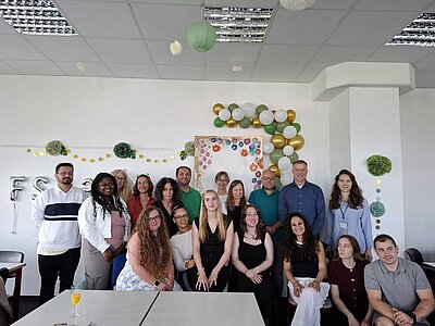 Eine Gruppe von Schüler*innen und Lehrer*innen haben sich für ein Foto in einem Klassenraum positioniert. Die Wand ist feierlich mit grünen, weißen und goldenen Luftballons und Pompons geschmückt. Man kann ganz hinten silberne Ballons erkennen, die die Buchstabenkombination FSK 23 bilden - ein Hinweis, dass es sich um die Klasse der Fremdsprachenkorrespondent*innen des Jahrgangs 2023 handelt.