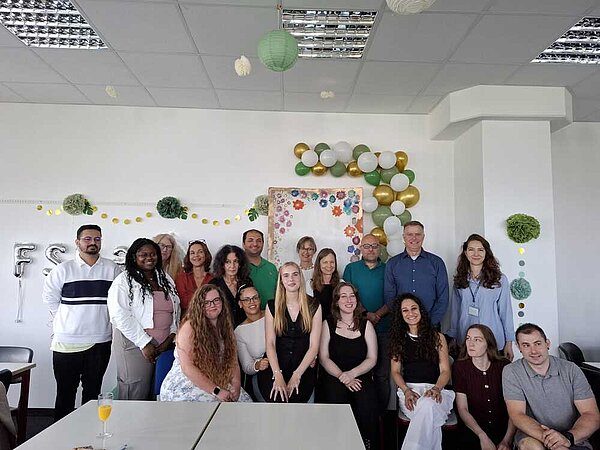 Eine Gruppe von Schüler*innen und Lehrer*innen haben sich für ein Foto in einem Klassenraum positioniert. Die Wand ist feierlich mit grünen, weißen und goldenen Luftballons und Pompons geschmückt. Man kann ganz hinten silberne Ballons erkennen, die die Buchstabenkombination FSK 23 bilden - ein Hinweis, dass es sich um die Klasse der Fremdsprachenkorrespondent*innen des Jahrgangs 2023 handelt.