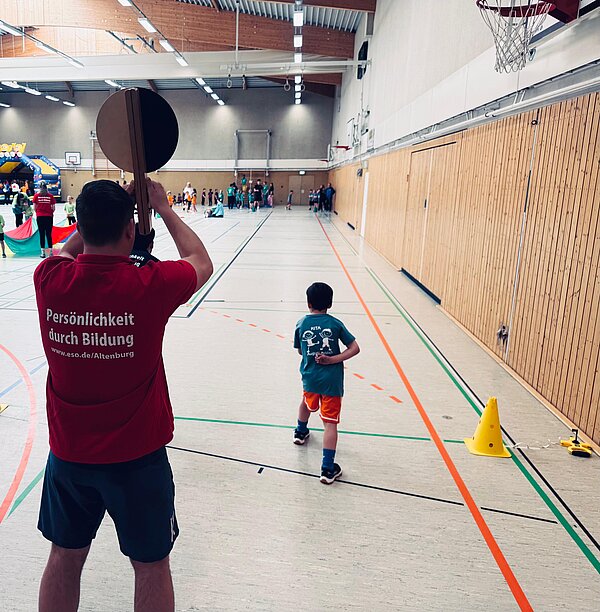 n einer Turnhalle läuft ein Kind auf einer markierten Bahn nach vorne, während eine betreuende Person im Vordergrund ein rundes Schild hochhält; im Hintergrund sind weitere Kinder und Gruppenaktivitäten zu sehen.