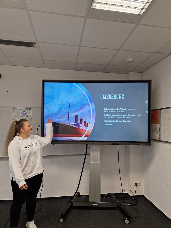 Eine Schülerin zeigt auf eine Präsentationsfolie mit dem Titel „Gliederung“ zum Thema Titanic. Auf dem Bildschirm ist ein Bild des Schiffs zu sehen, sowie die geplanten Themenpunkte der Präsentation.