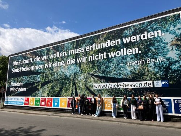 Eine große Plakatwand mit einem Zitat von Joseph Beuys, das besagt: "Die Zukunft, die wir wollen, muss erfunden werden. Sonst bekommen wir eine, die wir nicht wollen." Im Vordergrund steht eine Gruppe von Menschen, die vor dem Plakat posieren. Das Banner wirbt für das Museum Wiesbaden und die Website der 17 Ziele für nachhaltige Entwicklung.