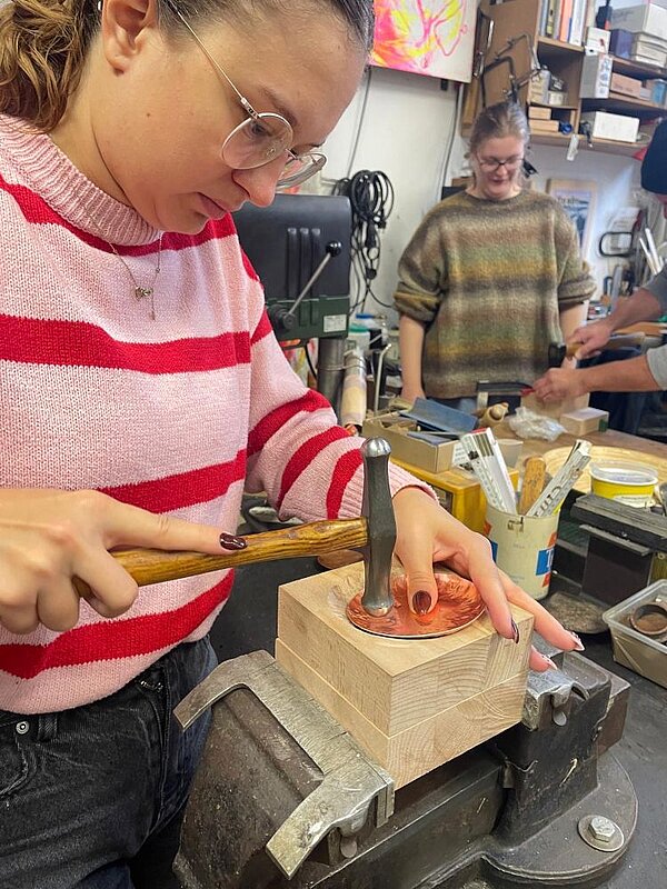 In einer Werkstatt schlägt eine Person mit rotgestreiftem Pullover und Brille mit einem Hammer auf ein Metallobjekt, das auf einem Holzblock liegt und mit der Hand festgehalten wird. Im Hintergrund arbeitet eine weitere Person in einem bunten Pullover. Die Umgebung ist mit Werkzeugen, Materialien und Regalen gefüllt.