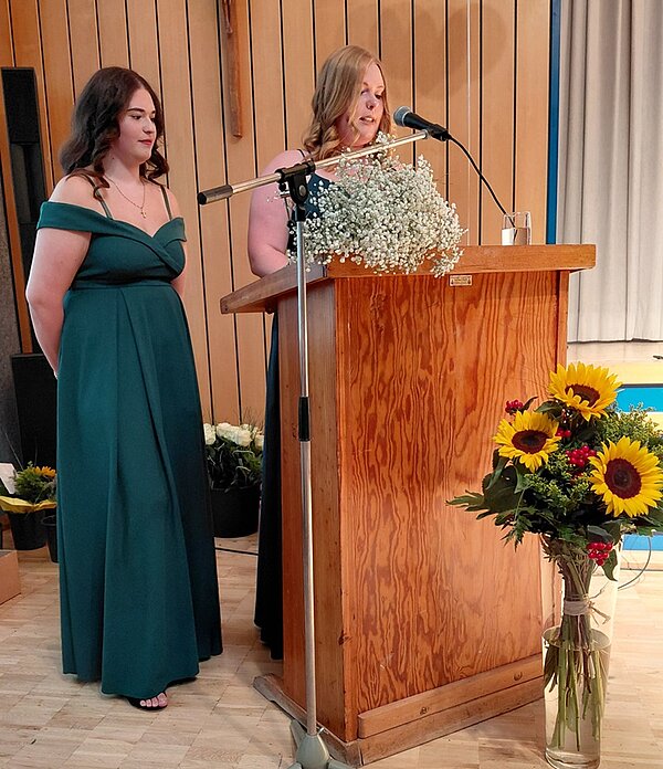 Zwei Absolventinnen in festlichen langen Kleidern stehen am Rednerpult. Eine spricht ins Mikrophon zum Publikum. Auf dem Pult liegen Blumen, auf dem Boden davor steht eine große gläserne Vase mit mehreren Sonnenblumen.