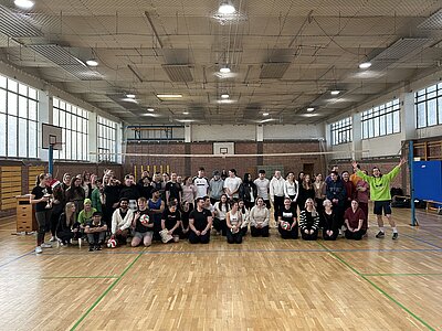 Gruppe aus Teilnehmenden des Volleyballturniers posiert in der Sporthalle, einige halten Volleybälle, vorne springt eine Person im neongrünen Shirt begeistert in die Luft.