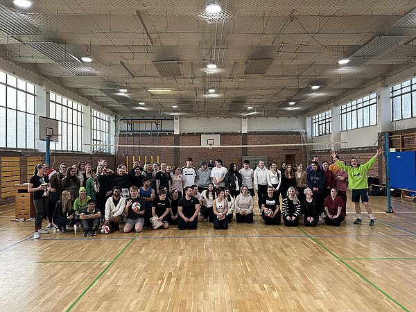 Gruppe aus Teilnehmenden des Volleyballturniers posiert in der Sporthalle, einige halten Volleybälle, vorne springt eine Person im neongrünen Shirt begeistert in die Luft.